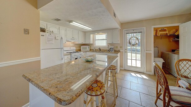 A kitchen with white cabinets, a granite countertop, and a large island with bar stools. - 206 White Sandy-pet Friendly (Panama City Beach)