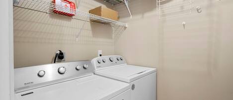 Ett tvättrum med en tvättmaskin och torktumlare från Maytag, en hylla med lådor och en röd behållare samt ett eluttag på väggen.