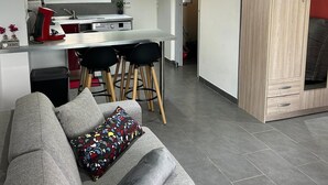 A modern living room with a grey sofa, a patterned throw pillow, and a rolled-up blanket. A kitchen with a white countertop and black chairs is visible in the background.