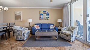 A living room with a blue sofa, two patterned armchairs, a coffee table, a ceiling fan, a dining table with chairs, and a large window with curtains. - Emerald Beach 1430 (Panama City Beach)