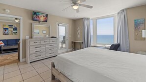 A bedroom with a large bed, a ceiling fan, a window with a view of the sea, a dresser, a mirror, and a TV. - Emerald Beach 1430 (Panama City Beach)