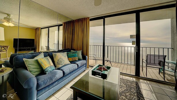 A living room with a blue sofa, a glass coffee table, and a balcony with a view of the ocean. - Edgewater 1008-3 (Panama City Beach)