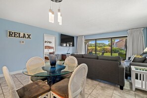 A modern living room with a glass dining table, wicker chairs, a grey sofa, and a TV mounted on the wall. - Edgewater Villa 1507 (Panama City Beach)