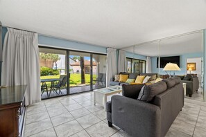 Een ruime woonkamer met een grote schuifpui naar een terras, een grijze zithoek, een klein witte salontafel en een lamp.