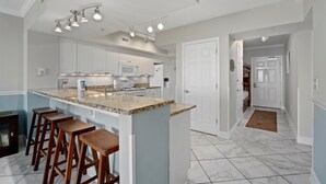 A modern kitchen with white cabinets, a granite countertop, and a bar stool area. - Emerald Beach 2325 (Panama City Beach)