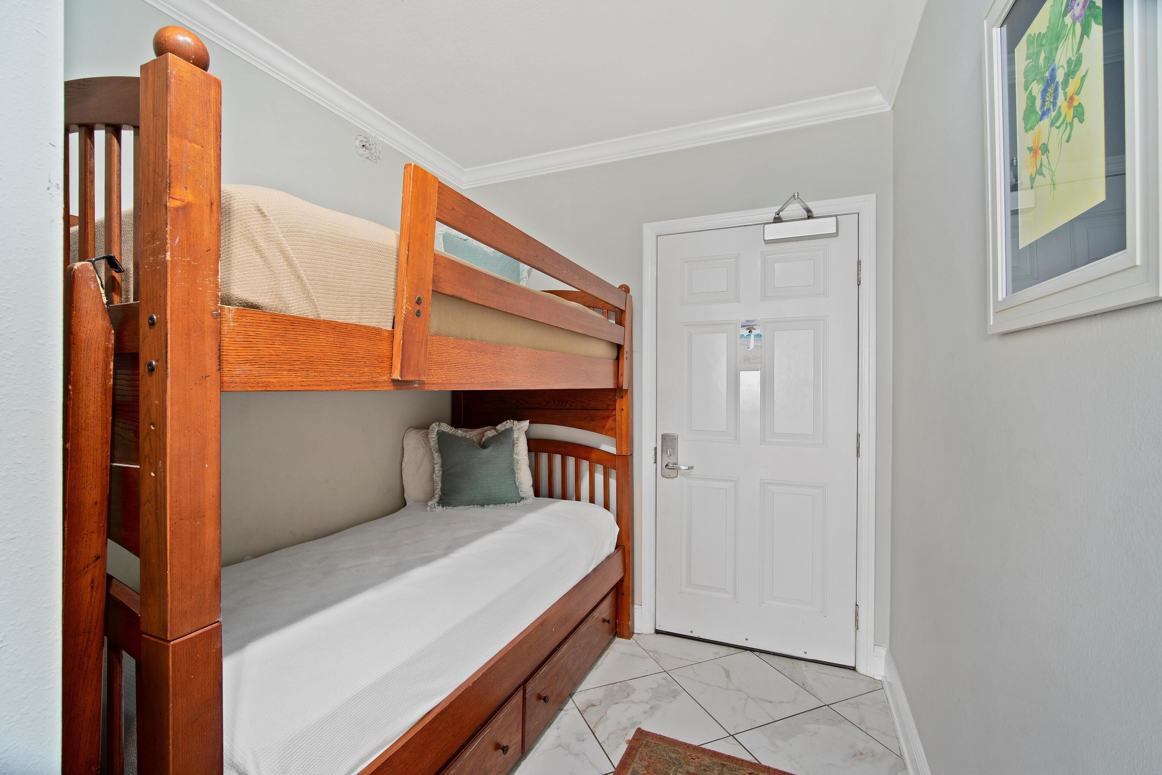 Una cama litera con estructura de madera, un colchón blanco y una sola almohada. Hay una puerta blanca con manija y un pequeño marco de fotos en la pared.