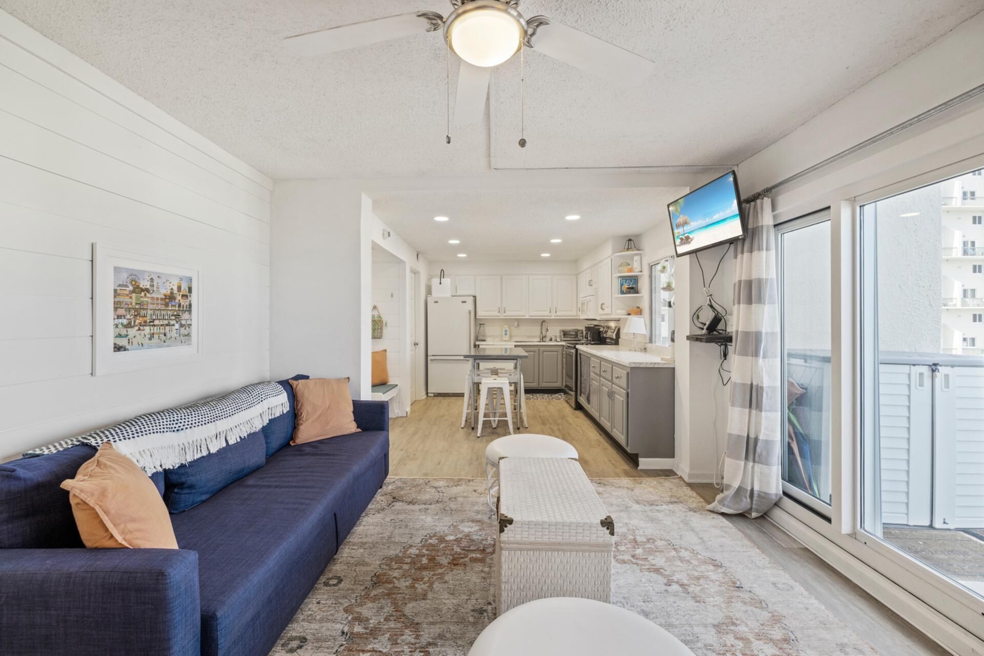 A modern living room with a sofa, a small table, and a bench. There is a kitchen area with cabinets and a television.