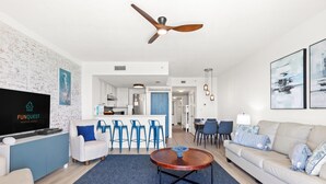 A modern living room with a flat-screen TV, a ceiling fan, a dining area with blue chairs, and a sofa with patterned cushions. - Seychelles 1006 (Panama City Beach)