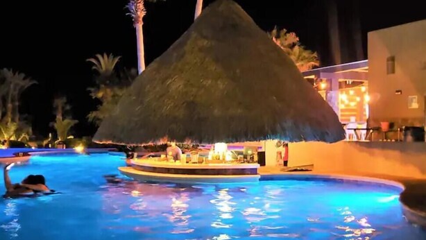 A swimming pool with a thatched-roof structure, palm trees, and a building with lights.
