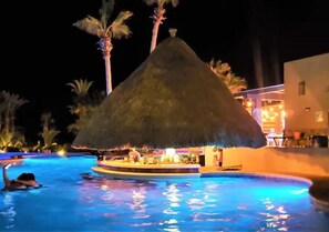 Une piscine dotée d’une structure à toit de chaume, de palmiers et d’un bâtiment illuminé.