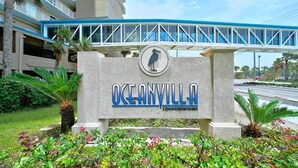 Oceanvilla-kerrostaloyhtiön kyltti, jossa on lintukuvio, jalankulkusilta ja runsasta vihreää kasvillisuutta.