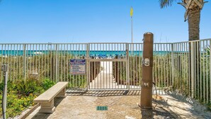 Une porte d’accès à la plage, agrémentée d’un panneau d’avertissement, d’un banc et d’un palmier.