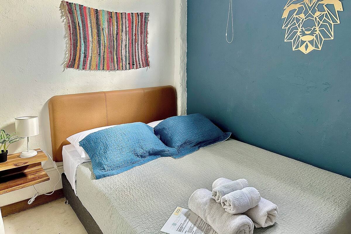 Un lit bien fait, avec des oreillers bleus et blancs, une tête de lit marron et une couverture blanche. Un tableau mural avec des rayures multicolores et une œuvre d’art géométrique ornent le mur.