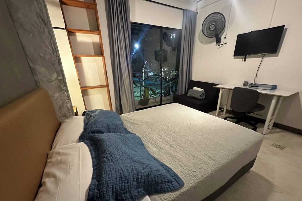 Un lit bien fait, recouvert d’une couverture bleue, un bureau avec une chaise, un téléviseur fixé au mur et une fenêtre avec des rideaux.