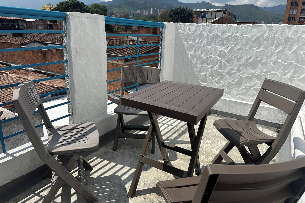 Un balcon avec une table et des chaises, un mur blanc et une vue sur des bâtiments et des montagnes en arrière-plan.