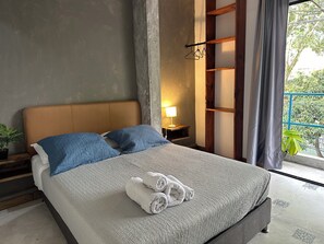 Een slaapkamer met een bed, kussens, een nachtkastje, een lamp en uitzicht op groen.
