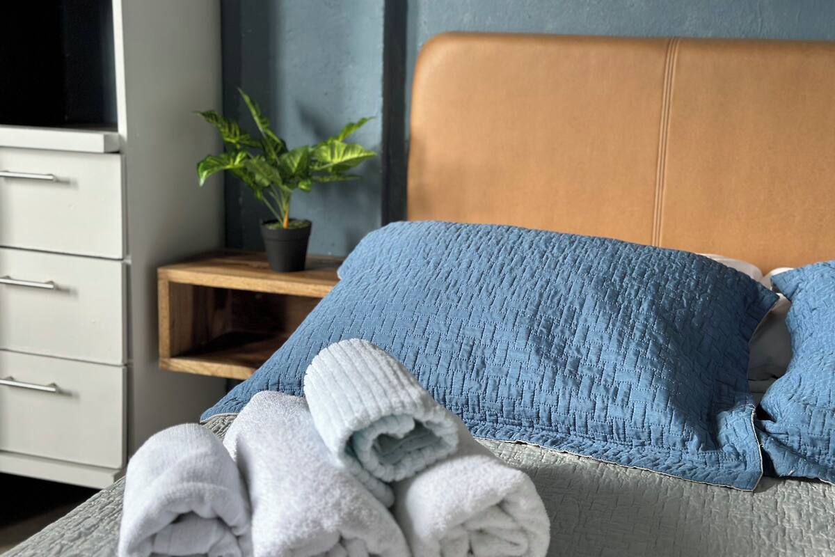 Un lit bien fait, avec des oreillers bleus et blancs, une serviette blanche roulée et une plante en pot posée sur une table de chevet en bois.