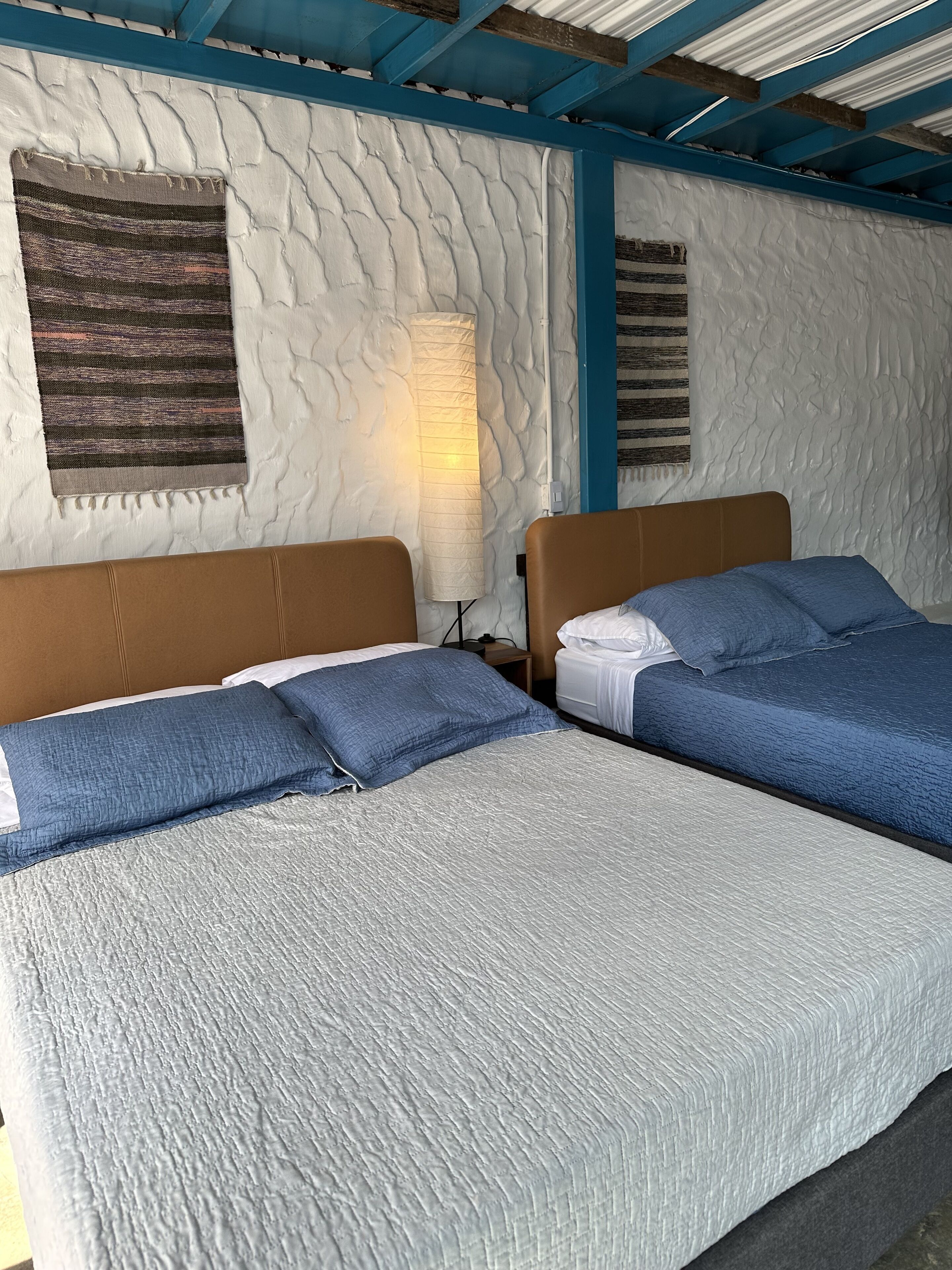Una habitación con dos camas, cada una con cabecera y almohadas, una pared con textura y un techo con vigas azules.