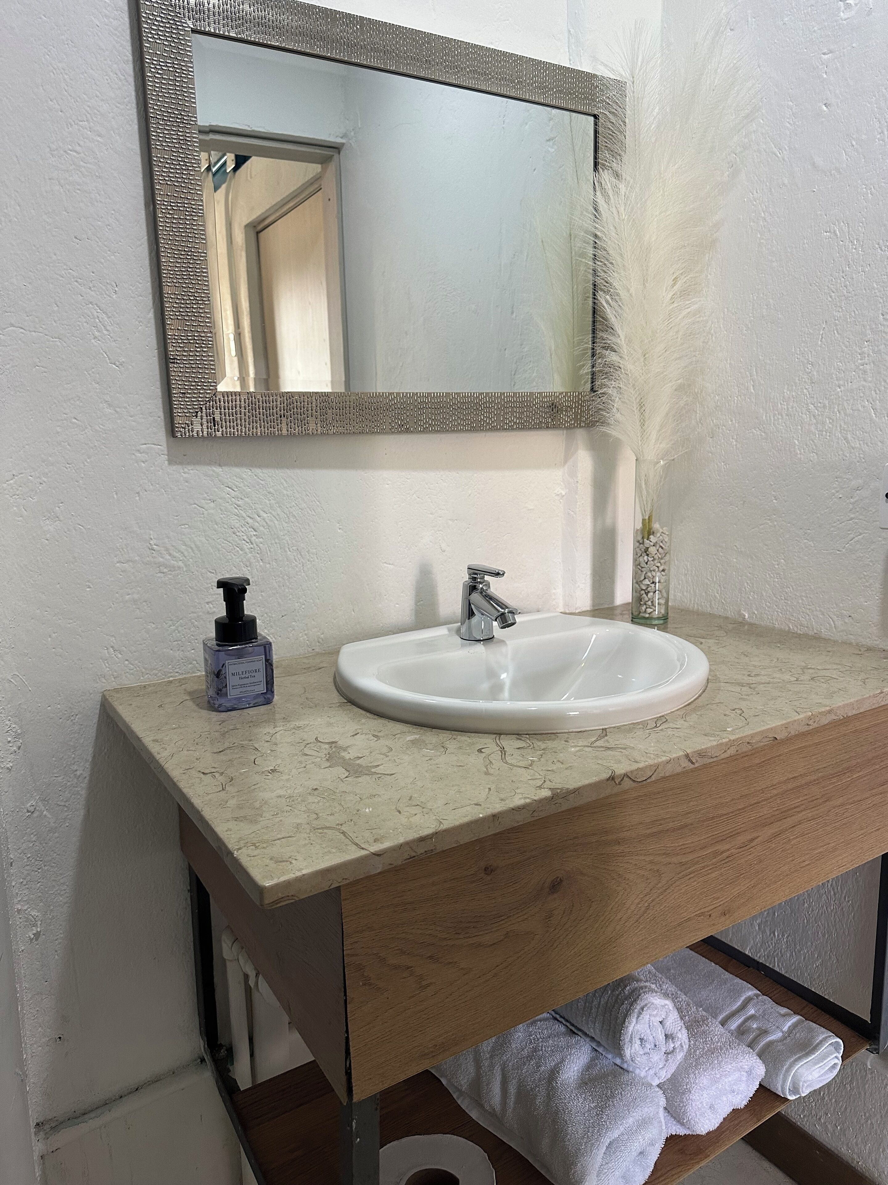 Casa de banho com lavatório branco, espelho com moldura texturizada, frasco de sabonete líquido e um vaso com penas brancas.