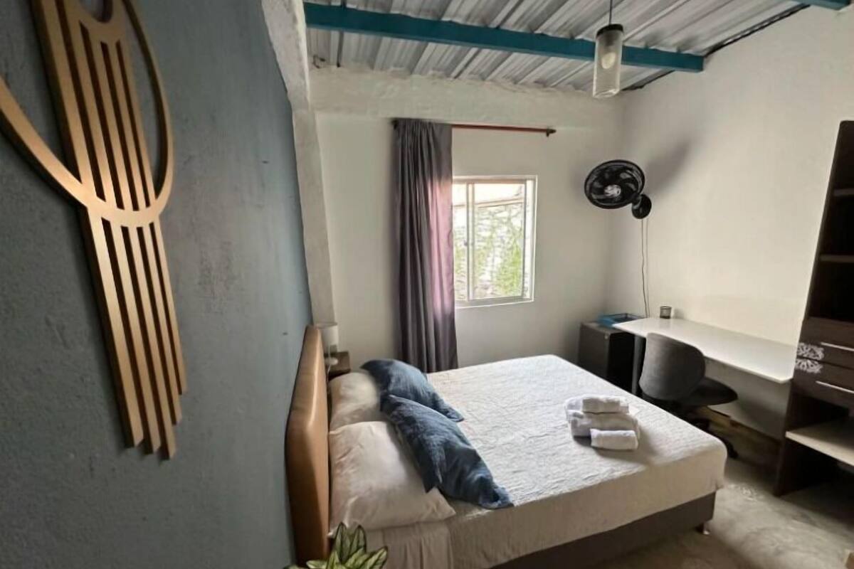Une chambre à coucher avec un lit, un bureau, un ventilateur et une fenêtre avec des rideaux.