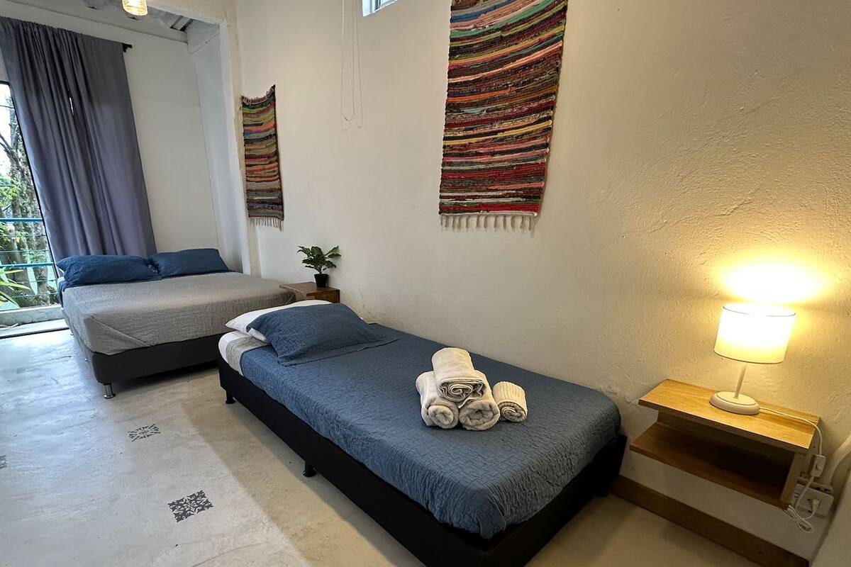 Une chambre avec un lit simple, un petit lit, une table de chevet avec une lampe et une fenêtre avec des rideaux.
