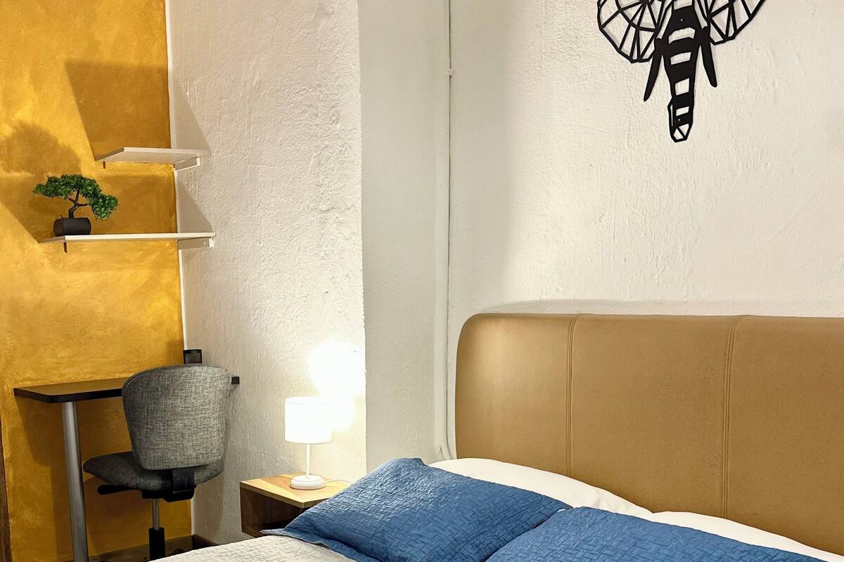 Une chambre d’hôtel comprenant un lit, un bureau, une chaise, une lampe et un élément décoratif mural.