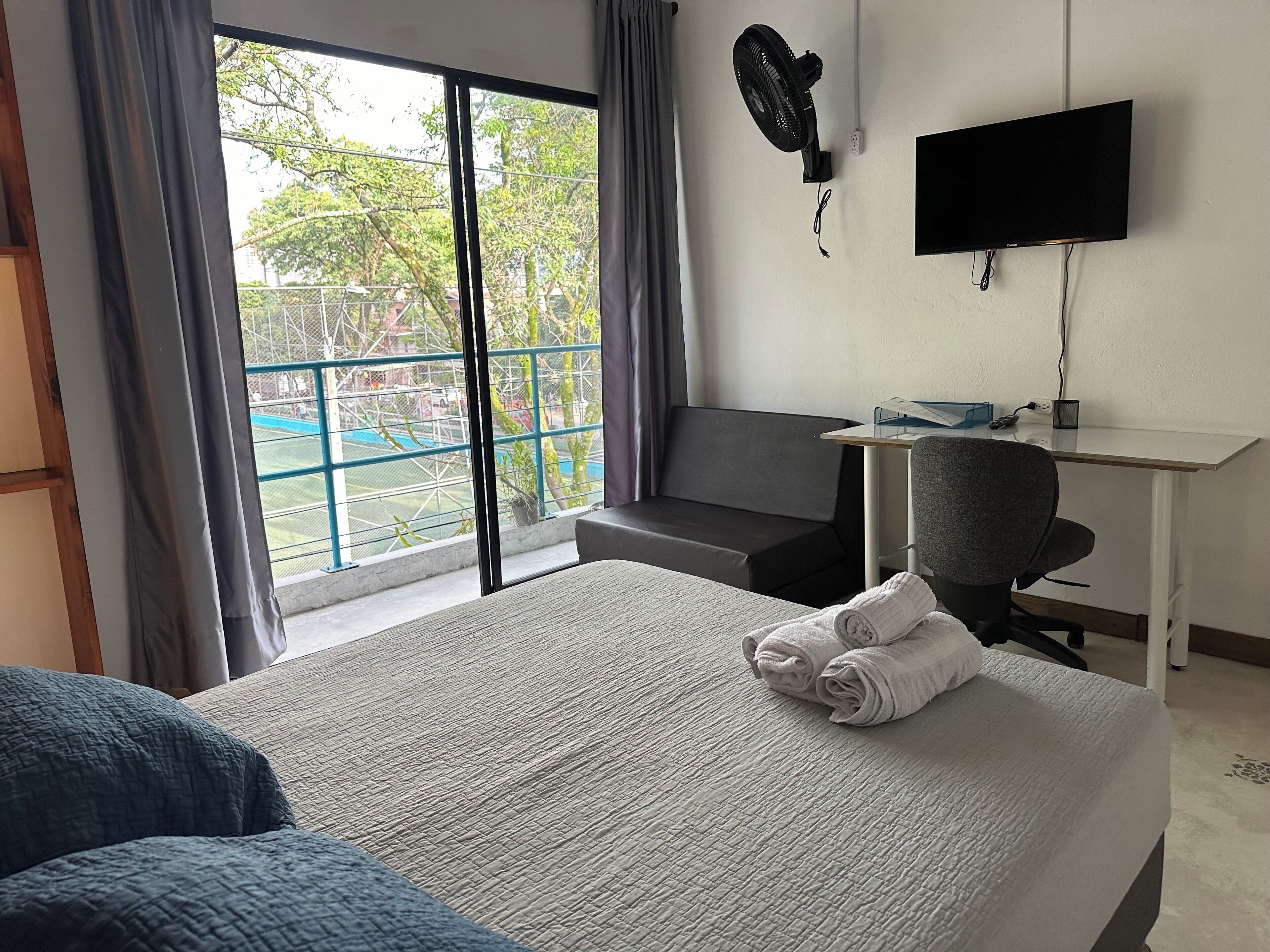 Habitación de hotel con una cama grande, un escritorio, una silla, un televisor y un balcón con vistas a zonas verdes.