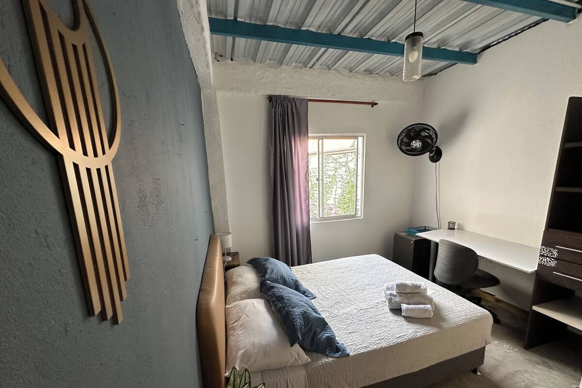 Une chambre à coucher avec un lit, un bureau, un ventilateur et une fenêtre.