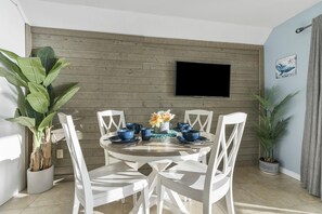 A dining area with a round table, white chairs, a TV mounted on the wall, and potted plants.