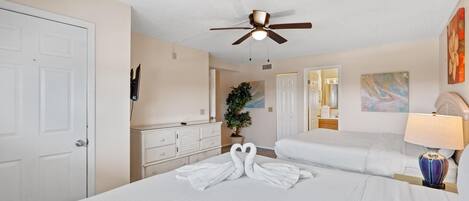 Habitación de hotel con una cama grande, un ventilador de techo, un aparador, una planta en maceta y un cuadro en la pared.