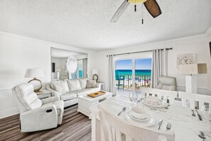 A spacious living room with a white sofa, a dining table set for two, and a large window offering a view of the ocean.