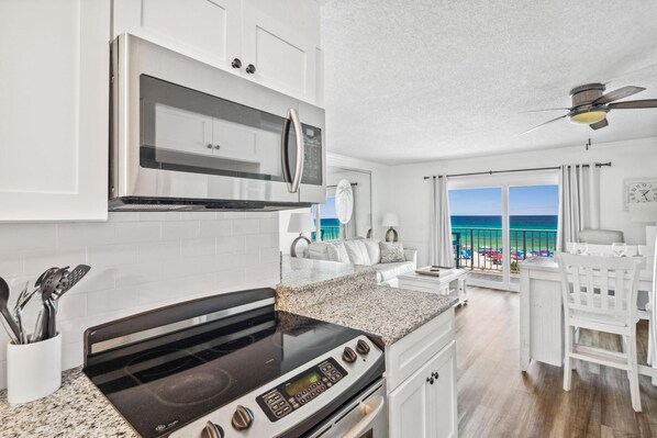 A modern kitchen with a stove, microwave, and white cabinets. There is a dining area with a white table and chairs, and a view of the ocean.