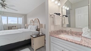 Quarto com cama, mesa de cabeceira e apoio para objetos. O banheiro possui pia, espelho e porta-toalhas.