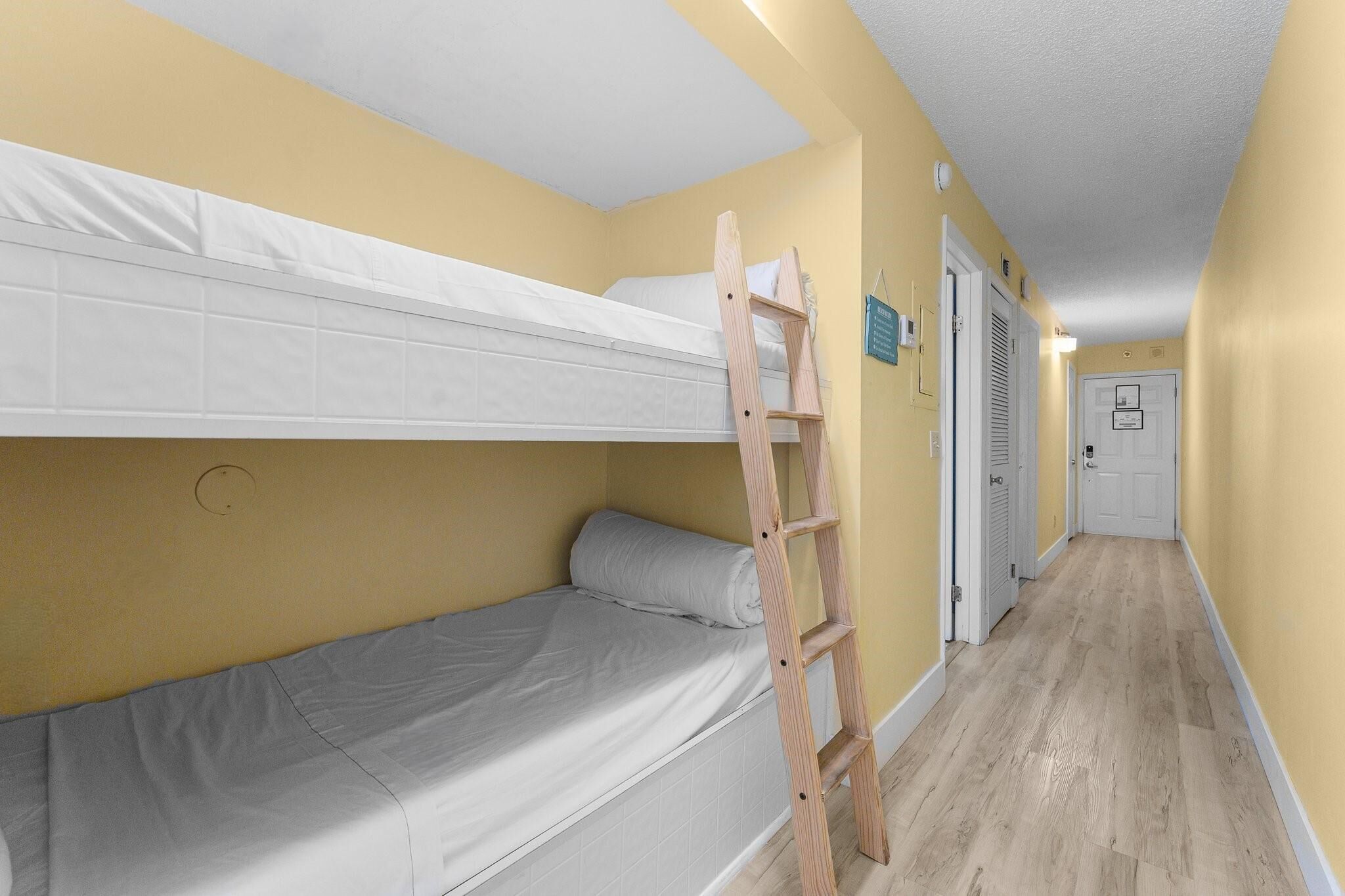 Una habitación con una cama litera, una escalera de madera, una puerta blanca y un pasillo con suelo de color claro.
