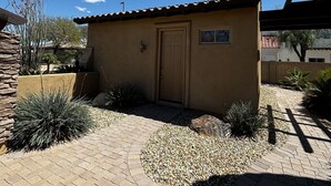A small, single-story building with a tiled roof and a door, surrounded by a gravel pathway and various plants.
