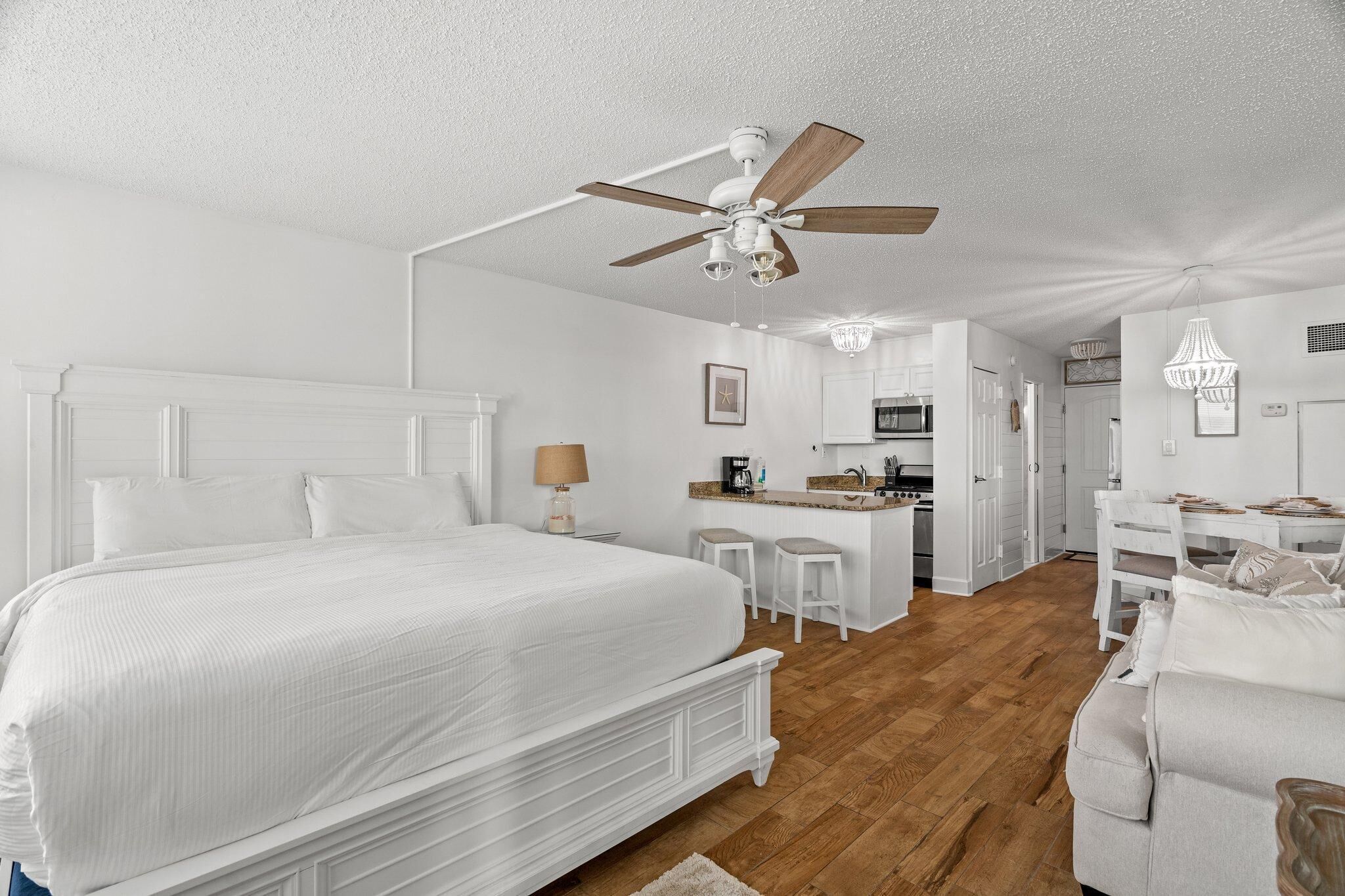 Une chambre avec un grand lit, un ventilateur de plafond et, au fond, un coin cuisine.