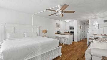 Une chambre avec un grand lit, un ventilateur de plafond et, au fond, un coin cuisine.