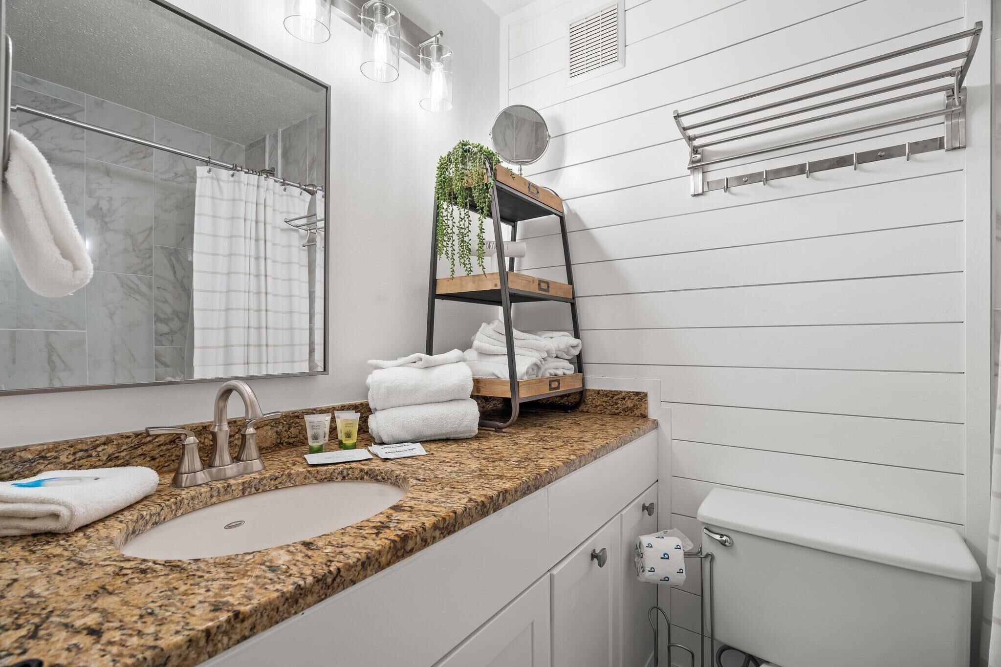 A bathroom with a granite countertop, a sink, a mirror, a towel rack, and a shelf with towels and a plant.