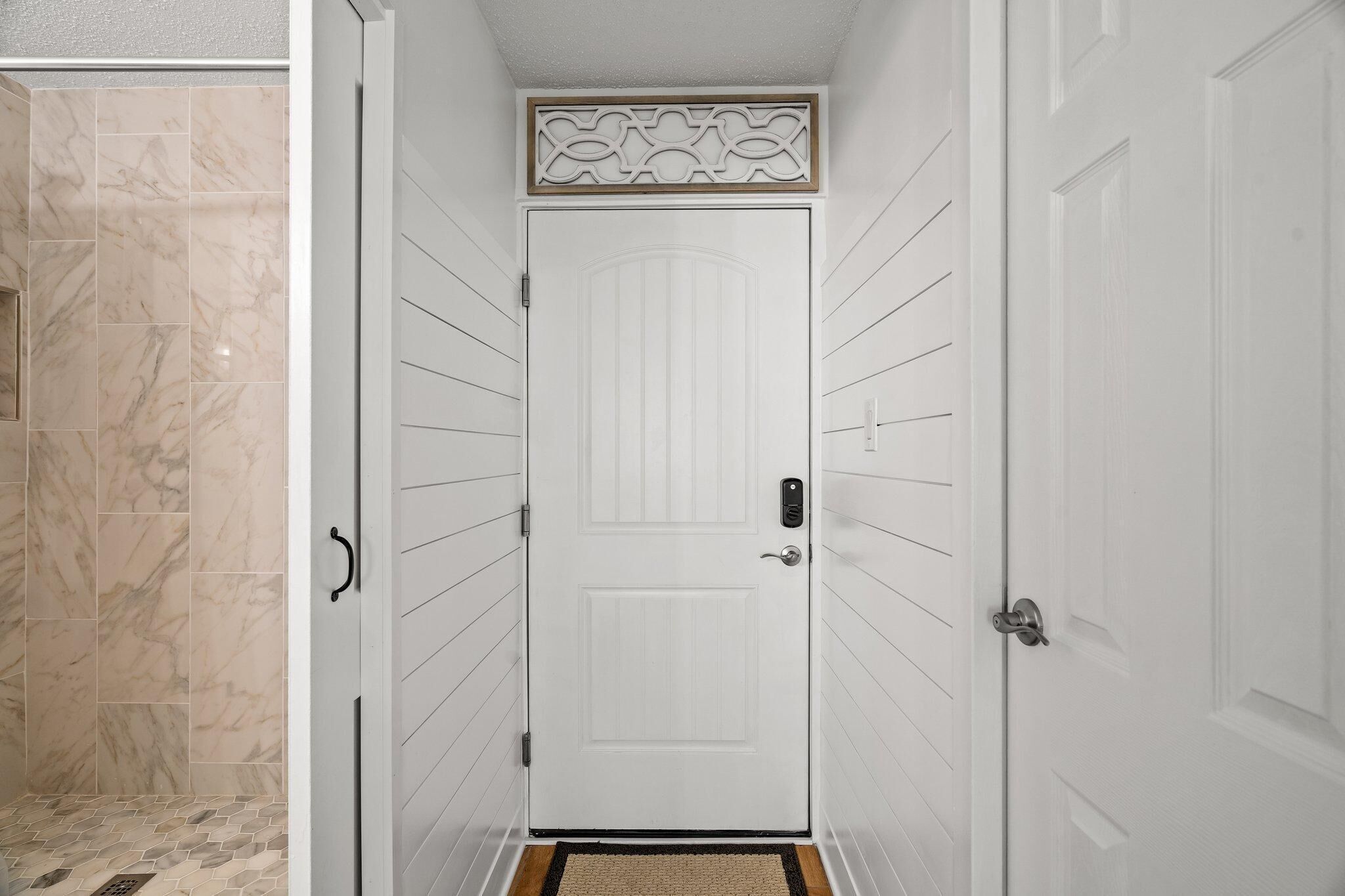 Una puerta blanca con un panel decorativo en la parte superior, una zona de ducha con azulejos color beige y una pequeña alfombra en el suelo.