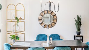Uma área de refeições com um espelho redondo, cadeiras estofadas em azul, uma mesa de madeira e um lustre.