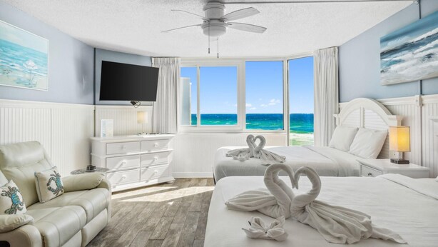 A hotel room with a large window offering a view of the ocean, a bed with two swan-shaped towels, a white sofa, a flat-screen TV, and a ceiling fan. - Top Of The Gulf 613 (Panama City Beach)