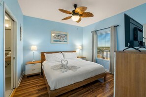 A bedroom with a wooden bed, two bedside lamps, a ceiling fan, a painting, a TV, and a window with curtains. - Splash 804e (Panama City Beach)