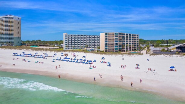 Ein Strandhotel mit mehreren Gebäuden, einem Sandstrand und Sonnenschirmen, und einem strahlend blauen Himmel.
