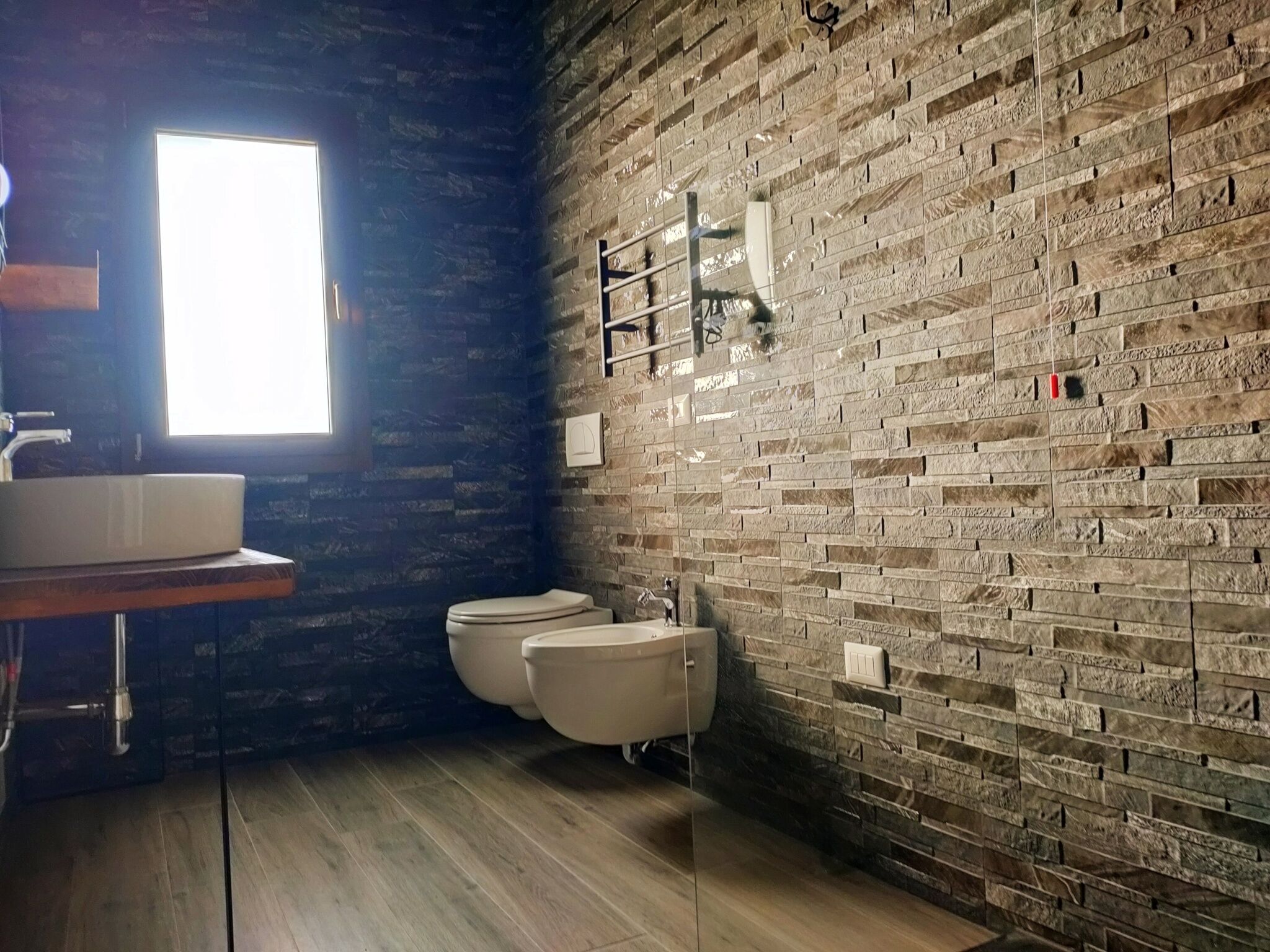 Ein Badezimmer mit rustikaler Ziegelwand, Holzboden, Waschbecken in Weiß und zwei Toiletten.
