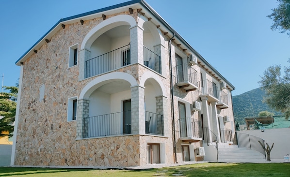 A two-story stone building with balconies, a stone facade, and a paved driveway leading to a gate.
