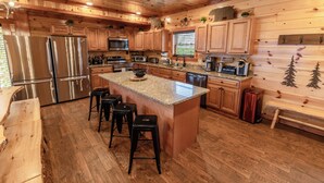 Interior de una cabaña de madera con cocina equipada, que incluye electrodomésticos de acero inoxidable, una isla con encimera de granito y taburetes de bar de madera.