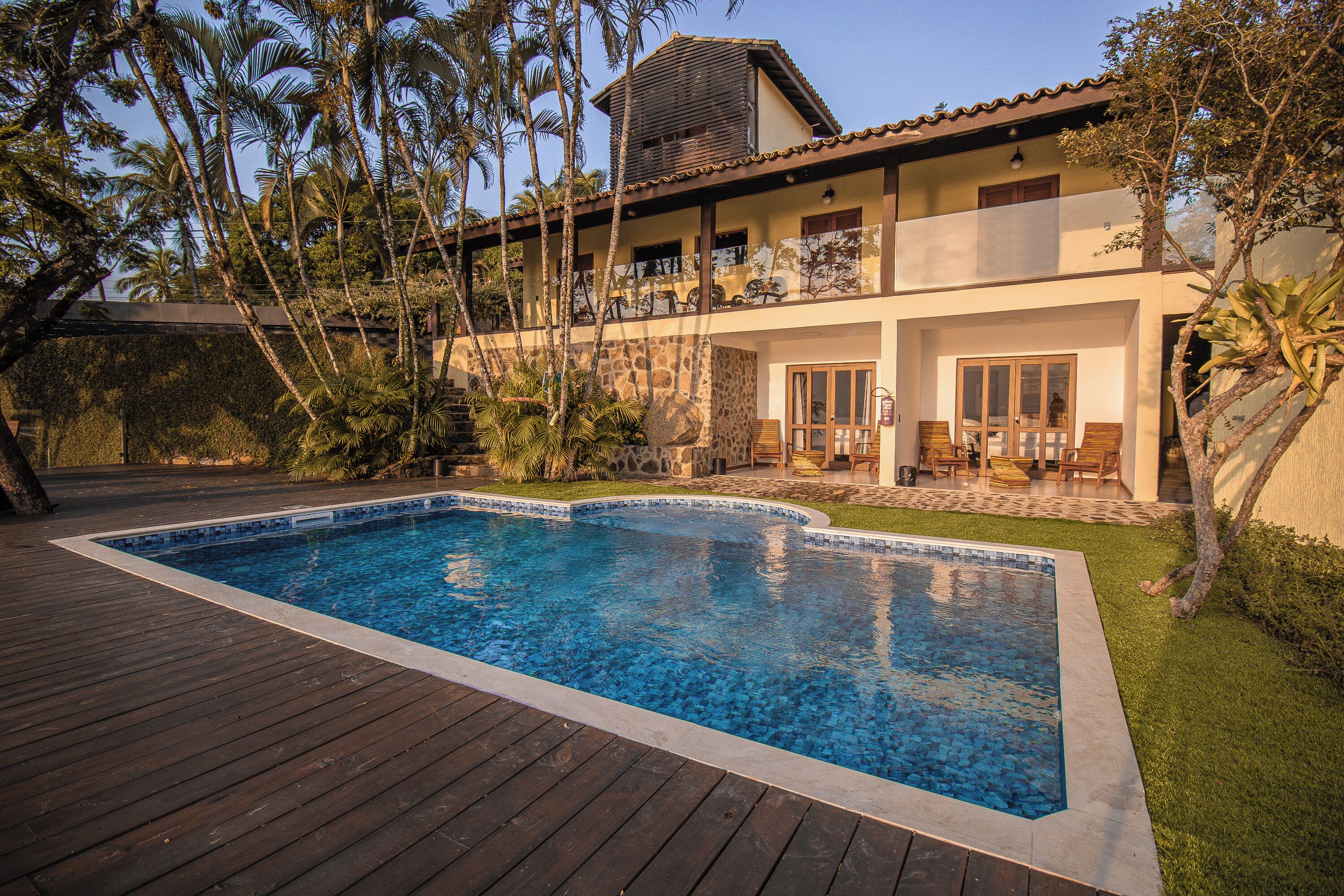 Une piscine dotée d’une terrasse en bois, entourée de palmiers et bordée d’un immeuble à deux étages avec des balcons.