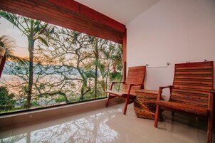 Una habitación con un ventanal grande que da a un paisaje tropical, dos sillas de madera y una pequeña mesa.