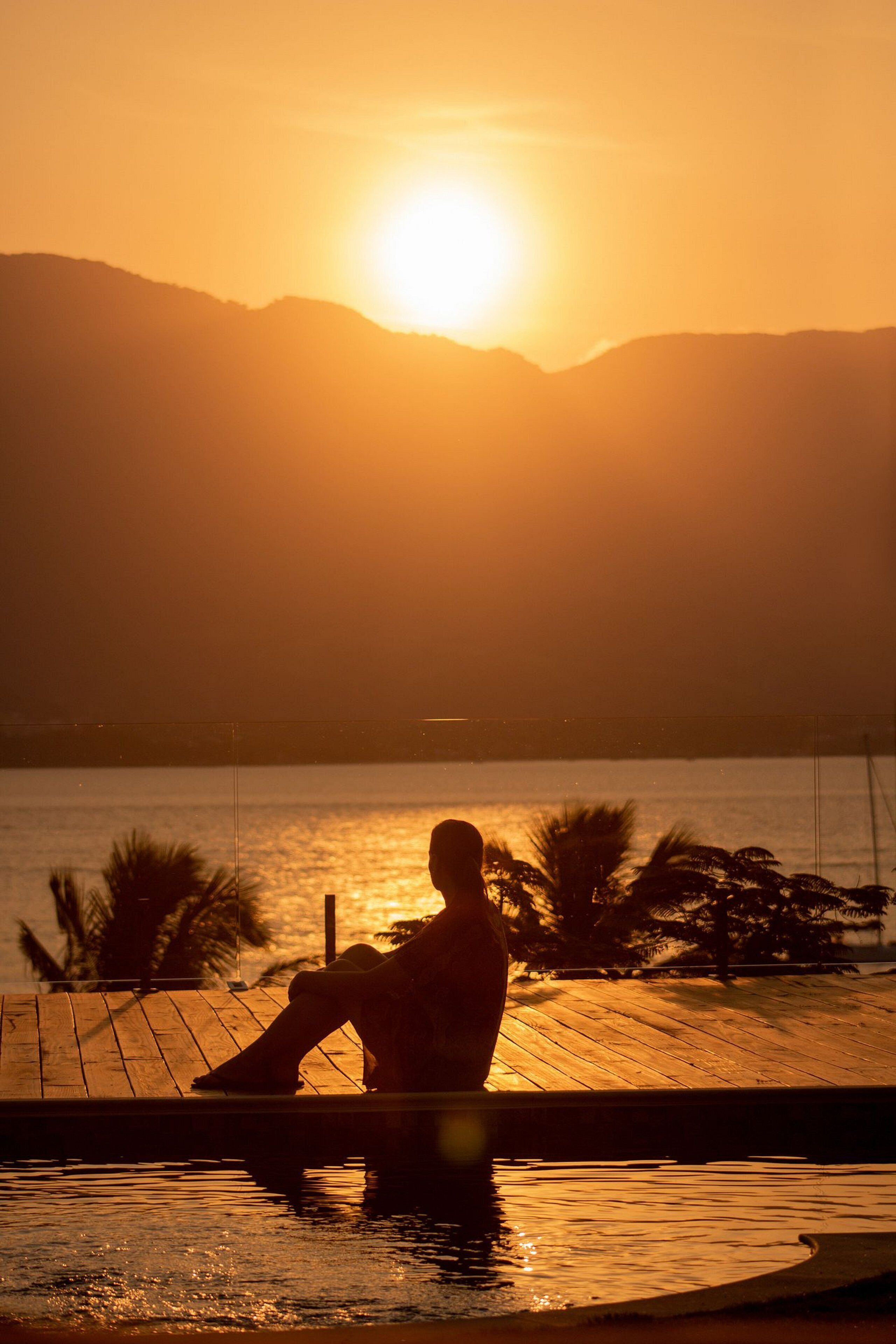 Uma pessoa sentada à beira da piscina durante o pôr do sol, com montanhas ao fundo.