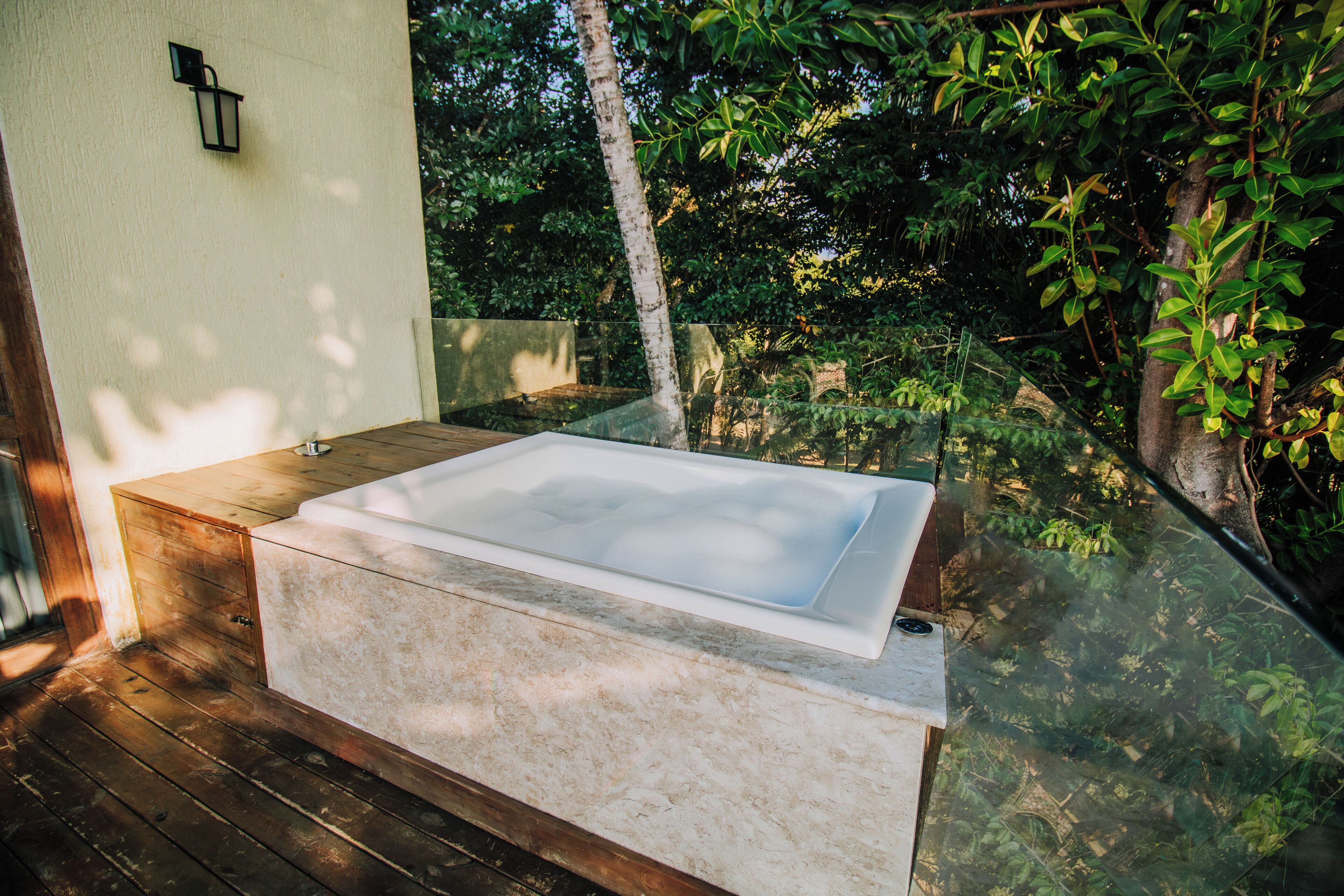 Un'area benessere con una grande vasca idromassaggio rettangolare su un pontile di legno, circondata da vegetazione lussureggiante e una struttura in vetro.